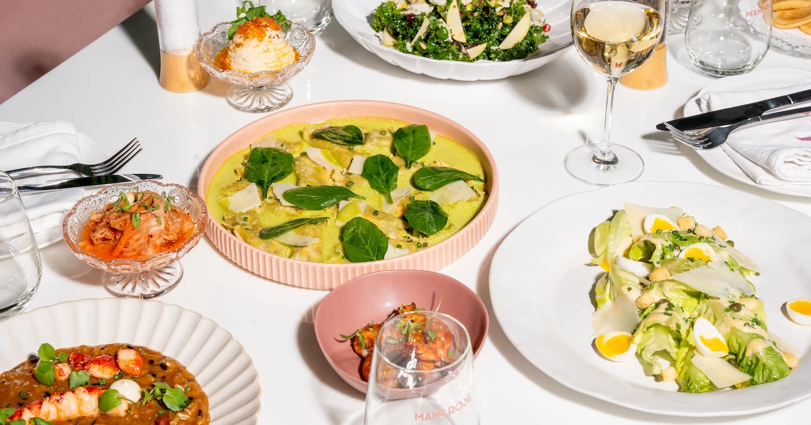 A table with a variety of dishes, including a green sauce ravioli dish, a fresh salad with eggs and parmesan, and small bowls of kimchi and other appetizers. Accompanied by glasses of white wine, creating an elegant and vibrant dining setting at Mami Rose restauant.