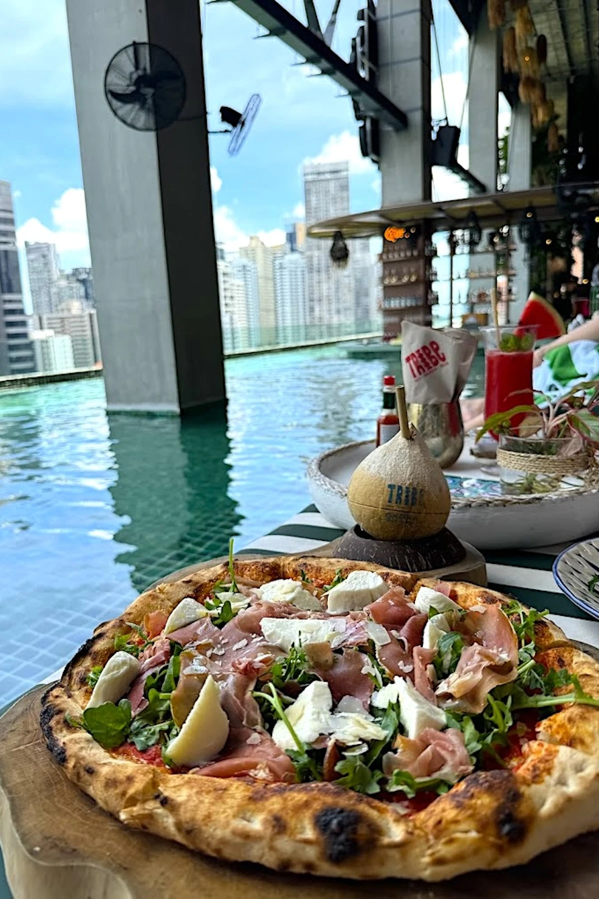 Pizza topped with arugula, ham, and cheese in front of an infinity pool with a city view and a coconut drink at Tribe Sky Beach Club