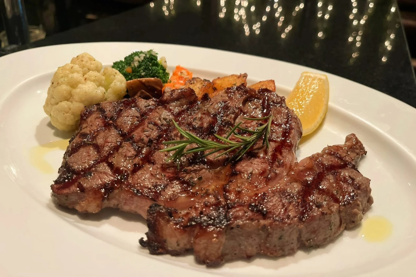 A perfectly grilled ribeye steak served with a side of roasted vegetables and a lemon wedge, garnished with a sprig of fresh rosemary. The dish showcases Antonio's attention to detail and love for premium ingredients.