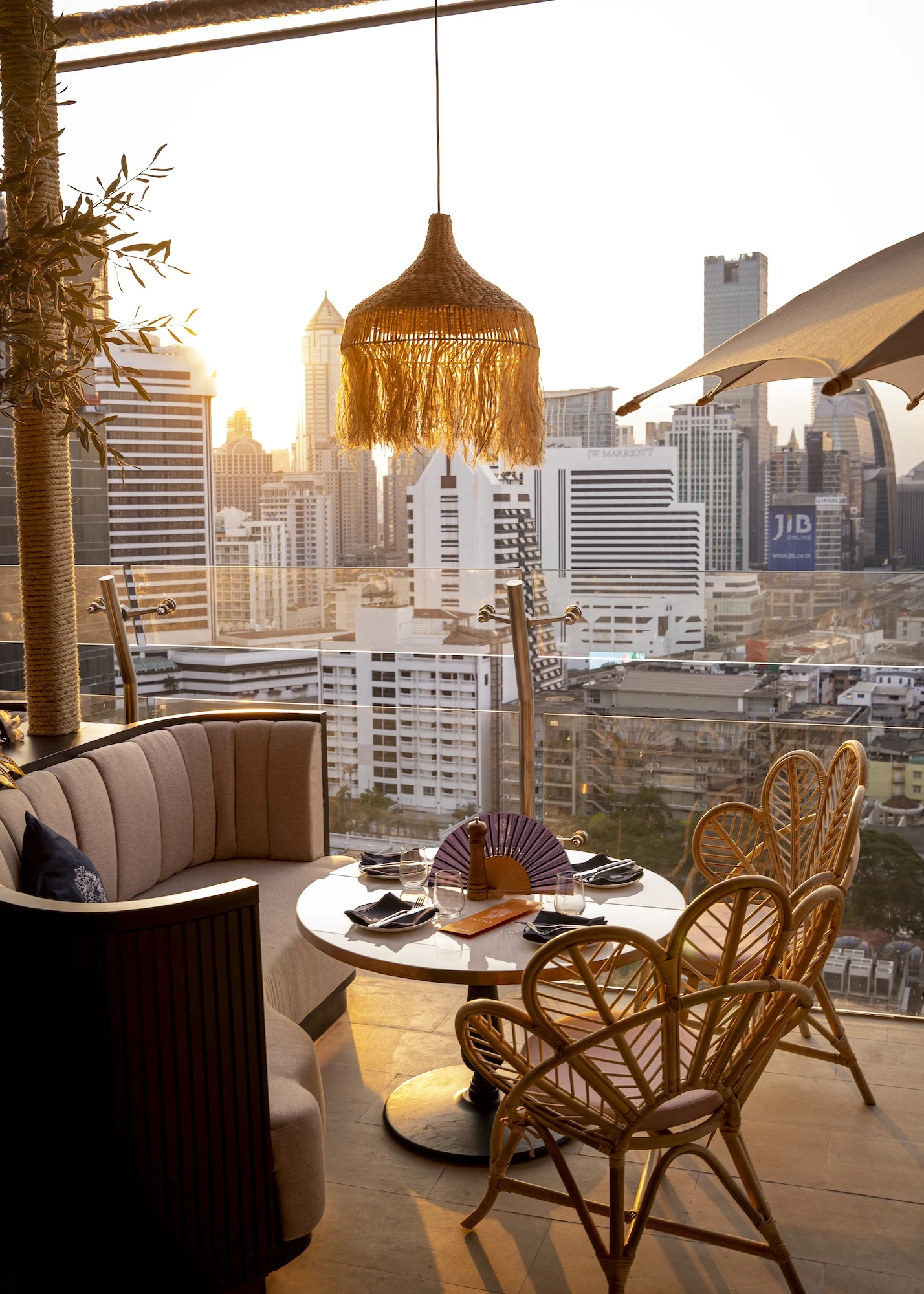 Outdoor terrace at Pastel Bangkok Rooftop Bar and Restaurant, featuring a cozy curved sofa and wicker chairs around a table, with a hanging wicker lamp and a stunning cityscape view at sunset