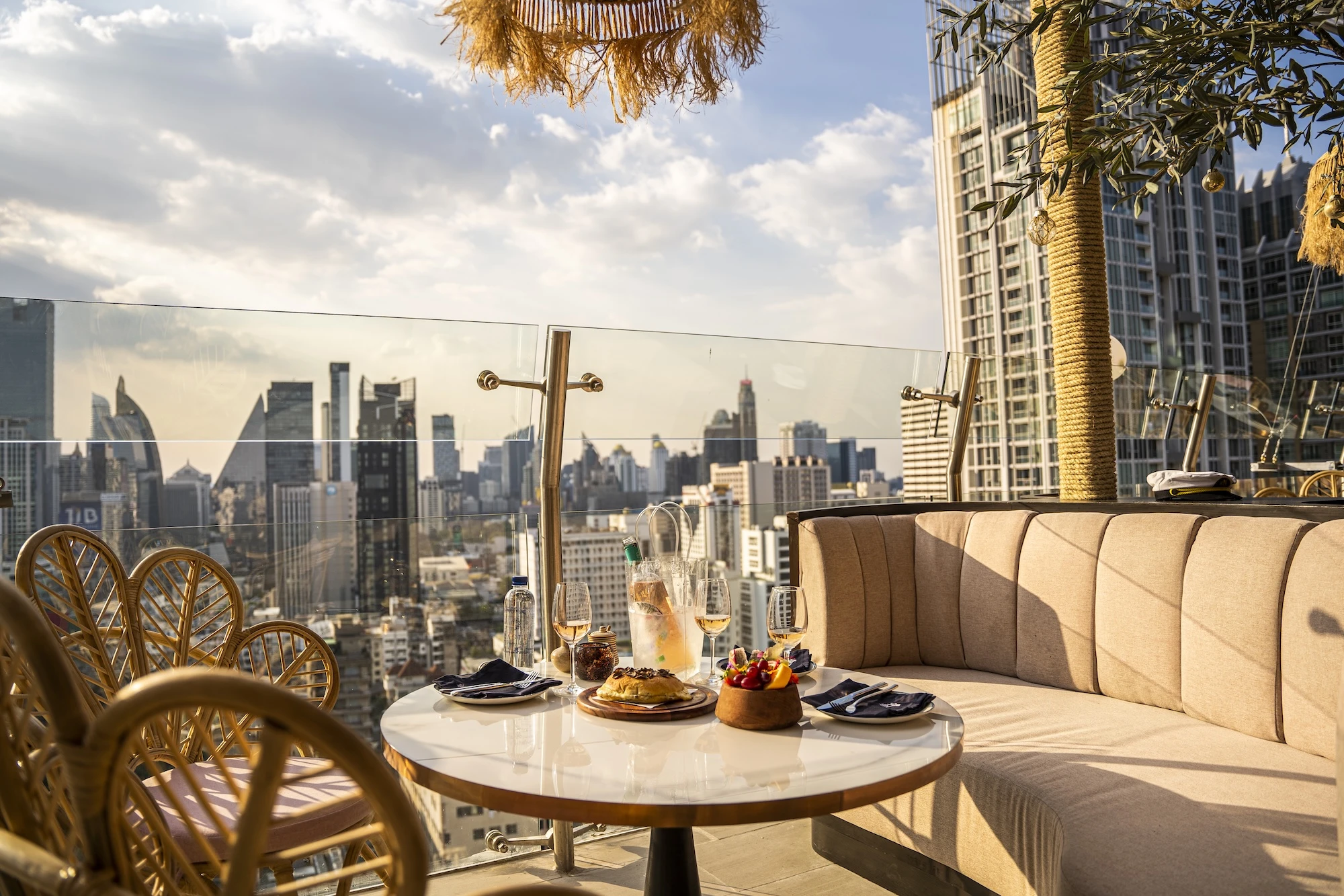 Terrace at Pastel Bangkok Rooftop Bar and Restaurant, featuring a curved sofa and wicker chairs around a table set with drinks and food, with a panoramic view of the Bangkok skyline in the background