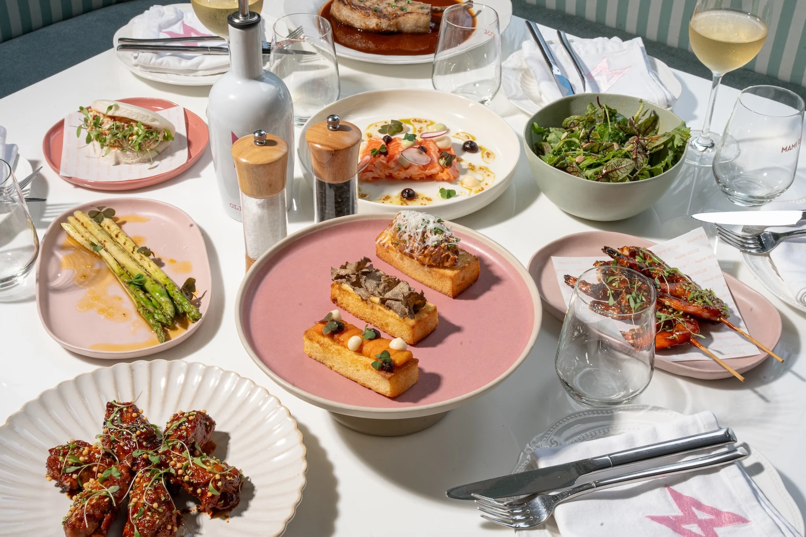 A table with assorted dishes: grilled shrimp skewers, a green salad, asparagus, truffle-topped toasts, glazed meat bites, salmon with garnish, and a soft bun sandwich. A refined and elegant setup with white wine at Mami Rose restaurant.